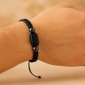 May include: Black braided bracelet featuring a rectangular black stone and two smaller, round, dark gray stones. The bracelet is adjustable with a sliding knot closure. A simple, elegant accessory.