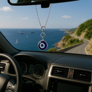 Peut inclure: Intérieur de voiture avec rétroviseur et tableau de bord. Une chaîne argentée pend du miroir, tenant un porte-bonheur en forme d'œil bleu et blanc. L'arrière-plan montre une route côtière et la mer.
