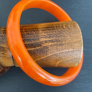 May include: Two bright orange bangles, likely made of resin or plastic, rest on a wooden form. The bangles have a smooth, glossy finish and a circular shape. The wood has a natural grain and a warm, brown tone. The background is a dark, neutral color.