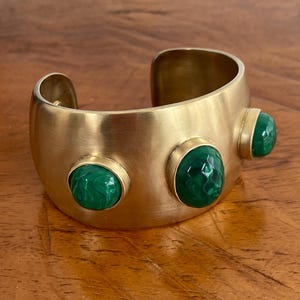 May include: A wide, gold-coloured cuff bracelet with three round, faceted, green malachite stones. The bracelet has a brushed metal finish and a simple, open design. The malachite stones are set in a bezel setting.