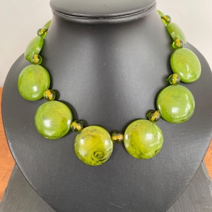 Puede incluir: Un collar con grandes cuentas de vidrio jaspeado en color verde oliva, intercaladas con cuentas más pequeñas, redondas y translúcidas que contienen una fina raya horizontal amarilla. Las cuentas están dispuestas en un patrón graduado.