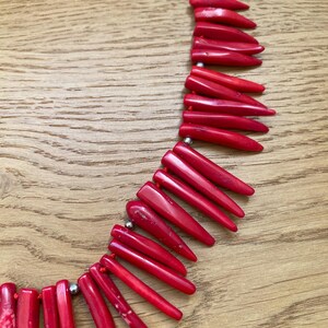 May include: A vibrant red necklace composed of elongated, pointed beads. The beads are strung together with a red cord and interspersed with small, silver-colored beads. The necklace is displayed on a wooden surface.