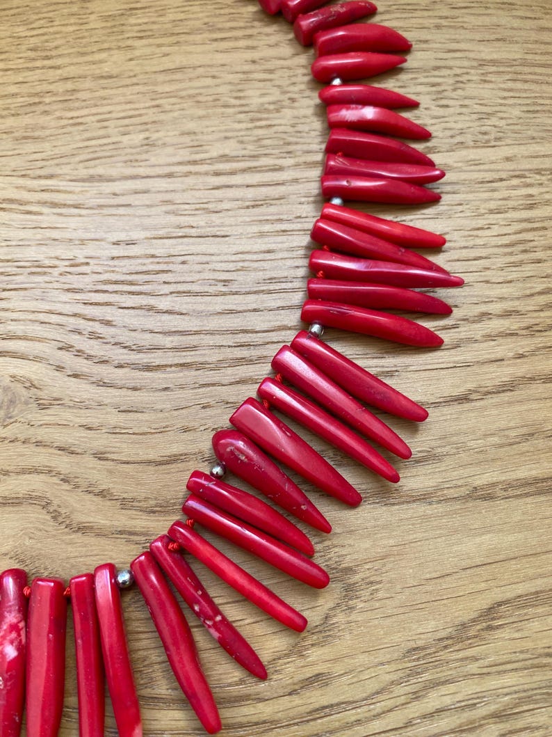 May include: A vibrant red necklace composed of elongated, pointed beads. The beads are strung together with a red cord and interspersed with small, silver-colored beads. The necklace is displayed on a wooden surface.