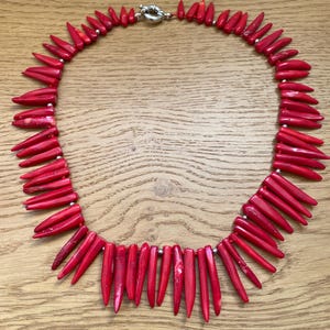 May include: A vibrant red necklace featuring numerous elongated, pointed beads. The beads are strung together with small silver-colored spacers and a silver clasp. The necklace is displayed on a light brown wooden surface.