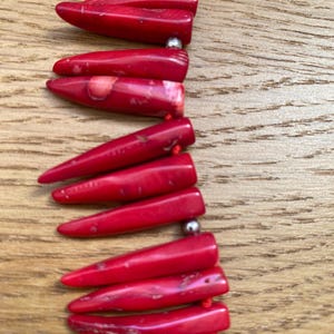 May include: A close-up of a red necklace with elongated, pointed beads. The beads are strung together with a red cord and separated by small, silver-colored beads. The necklace is displayed on a wooden surface.