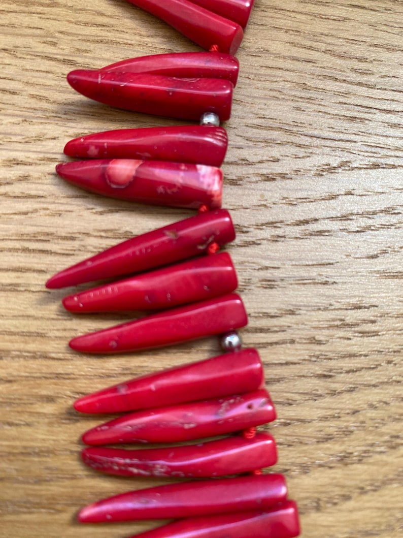 May include: A close-up of a red necklace with elongated, pointed beads. The beads are strung together with a red cord and separated by small, silver-colored beads. The necklace is displayed on a wooden surface.
