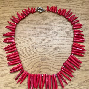 May include: A vibrant red necklace featuring numerous pointed, coral-like beads. The beads are strung together with small silver-colored beads and a silver clasp. The necklace is displayed on a light brown wooden surface.