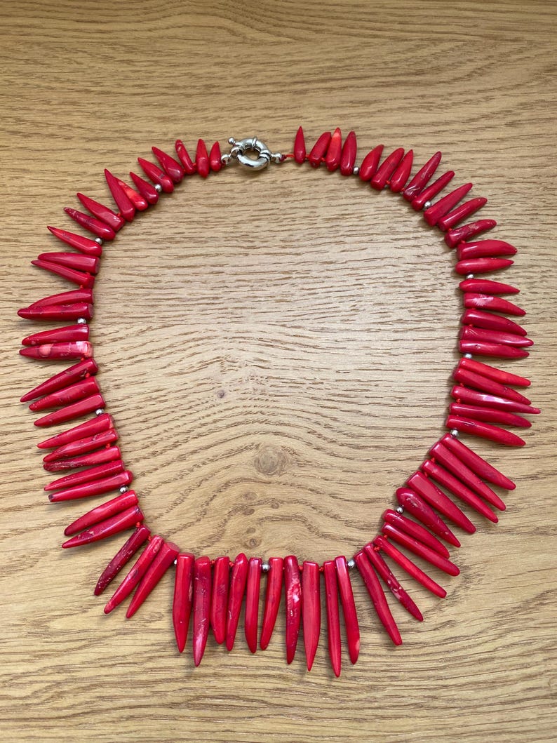 May include: A vibrant red necklace featuring numerous pointed, coral-like beads. The beads are strung together with small silver-colored beads and a silver clasp. The necklace is displayed on a light brown wooden surface.