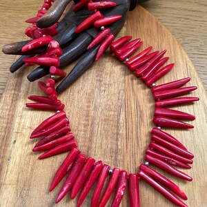 May include: A vibrant red coral necklace with a matching bracelet, displayed on a wooden hand. The necklace features elongated, pointed coral pieces strung together with silver-toned beads. The jewelry is set against a wooden background.