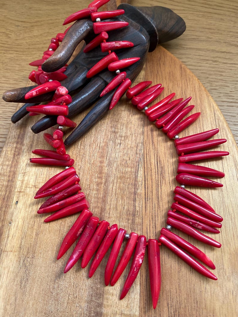 May include: A vibrant red coral necklace with a matching bracelet, displayed on a wooden hand. The necklace features elongated, pointed coral pieces strung together with silver-toned beads. The jewelry is set against a wooden background.