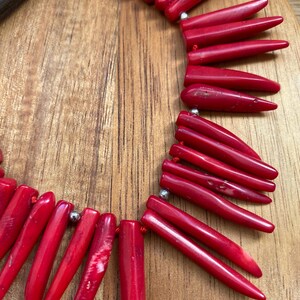 May include: A vibrant red necklace featuring numerous elongated, pointed beads. The beads are strung together with small silver-colored connectors. The necklace is displayed on a wooden surface, showcasing its color and texture.