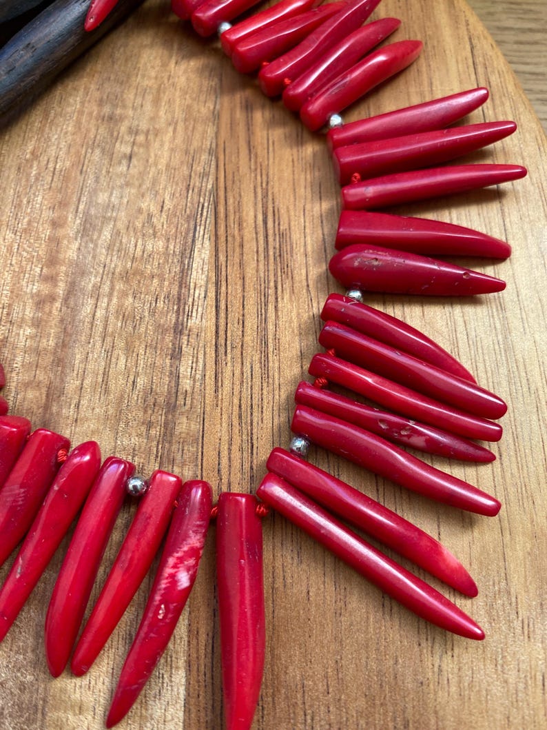 May include: A vibrant red necklace featuring numerous elongated, pointed beads. The beads are strung together with small silver-colored connectors. The necklace is displayed on a wooden surface, showcasing its color and texture.