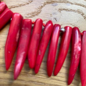 May include: A close-up of a red necklace with elongated, pointed beads. The beads are strung together with a red cord and small silver-colored beads. The necklace is set against a wooden background.