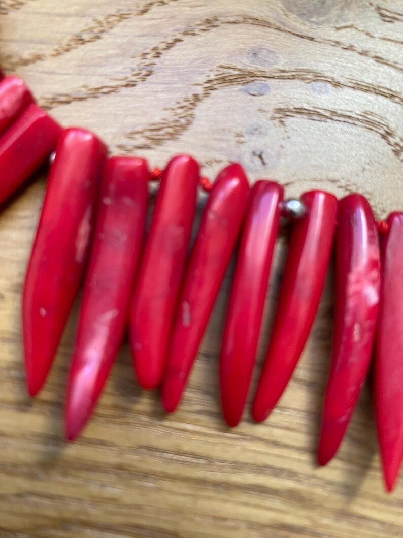 May include: A close-up of a red necklace with elongated, pointed beads. The beads are strung together with a red cord and small silver-colored beads. The necklace is set against a wooden background.