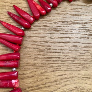 May include: A close-up of a red coral necklace with a silver clasp. The necklace features polished, elongated coral beads strung together. The beads are a vibrant red color and are arranged in a pattern. The background is a wooden surface.
