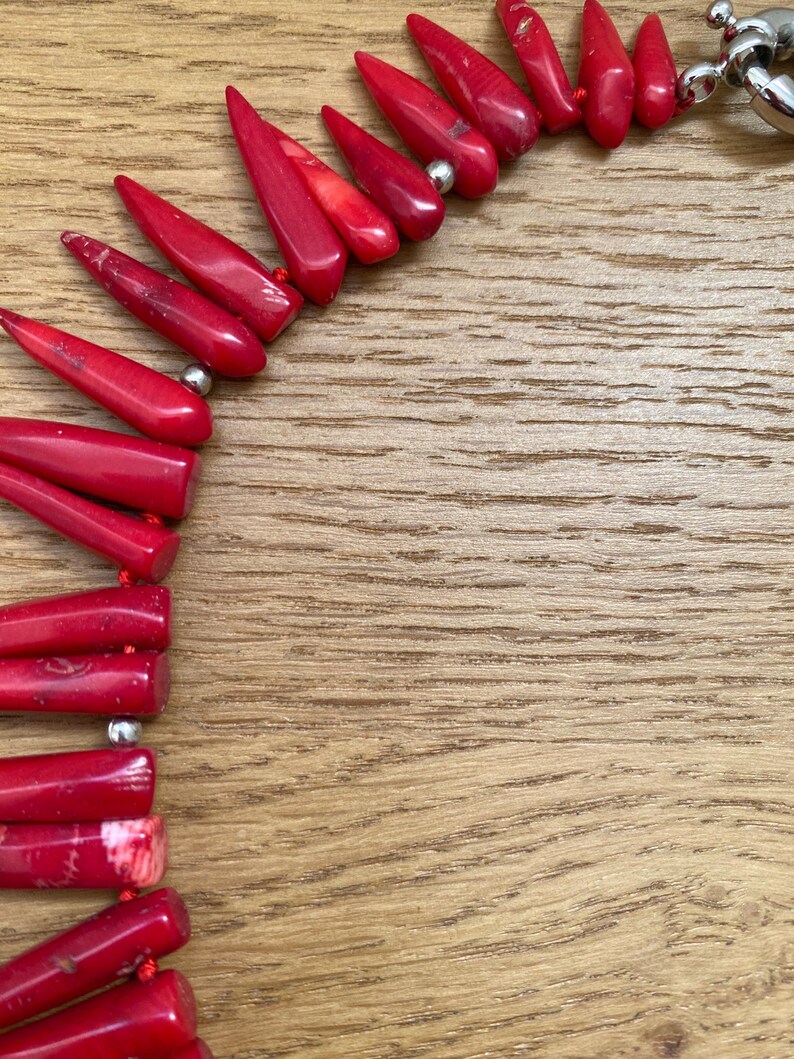 May include: A close-up of a red coral necklace with a silver clasp. The necklace features polished, elongated coral beads strung together. The beads are a vibrant red color and are arranged in a pattern. The background is a wooden surface.