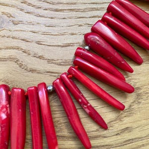 May include: A vibrant red necklace featuring elongated, pointed beads strung on a red cord. The beads have a glossy finish and are connected by small silver-colored beads. The necklace is displayed on a wooden surface.