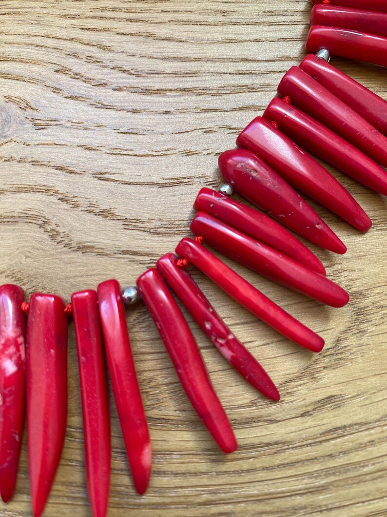 May include: A vibrant red necklace featuring elongated, pointed beads strung on a red cord. The beads have a glossy finish and are connected by small silver-colored beads. The necklace is displayed on a wooden surface.