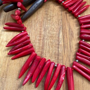 May include: A vibrant red coral necklace featuring elongated, pointed beads strung together with silver-colored accents. The necklace is displayed on a wooden surface, showcasing the natural texture and color of the coral.