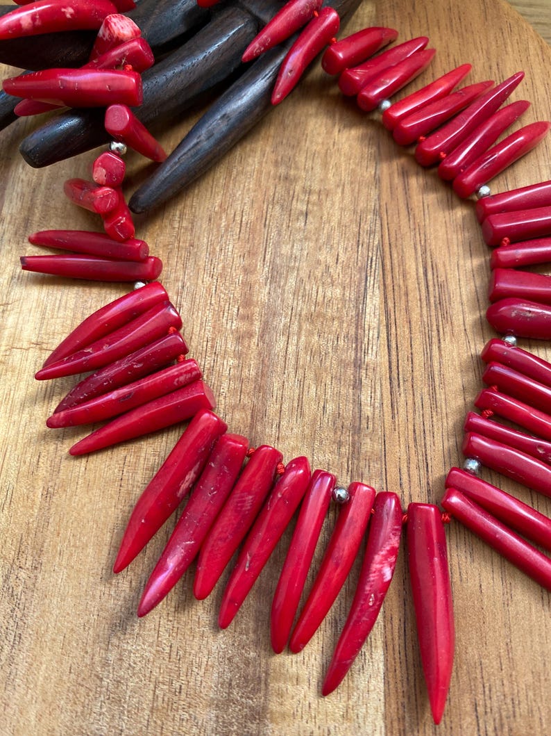 May include: A vibrant red coral necklace featuring elongated, pointed beads strung together with silver-colored accents. The necklace is displayed on a wooden surface, showcasing the natural texture and color of the coral.