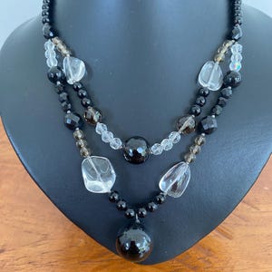 May include: A multi-strand necklace featuring a combination of smooth black spherical beads, faceted dark brown beads, and clear, irregular-shaped translucent stones. The design includes a large, polished black spherical pendant at the centre.