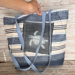 May include: A blue and white striped tote bag with light blue straps. The bag features a square patch with a white sea turtle design. The bag is held by a hand.