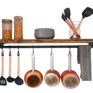 Puede incluir: Una estantería de madera rústica con una barandilla y ganchos de metal negro. La estantería contiene dos tarros de vidrio llenos de pasta, un cuenco y un portautensilios de cobre con espátulas de silicona negra. Cinco cacerolas de cobre cuelgan de los ganchos.