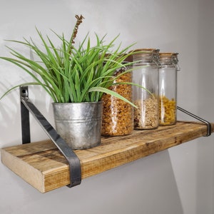 May include: A rustic wooden shelf with metal brackets, holding three glass jars filled with dried pasta and a potted green plant. The shelf is mounted on a white wall.