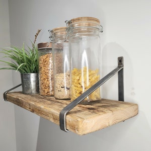 Könnte beinhalten: Ein rustikales Holzregal mit Metallkonsolen, das drei Glasbehälter mit Pasta und Müsli beherbergt. Das Regal ist an einer weißen Wand montiert.