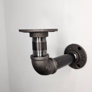 Puede incluir: Soporte de estante de tuber&iacute;a de metal negro con una brida en un extremo y un codo de 90 grados en el otro extremo. La tuber&iacute;a est&aacute; roscada y tiene un accesorio de color plateado.