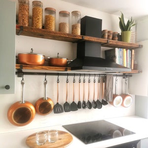 Puede incluir: Una cocina con una campana extractora negra y dos estantes de madera. Los estantes están llenos de frascos de pasta, granos y especias. Hay un estante de metal negro con ganchos debajo de los estantes, que sostiene ollas y sartenes de cobre.