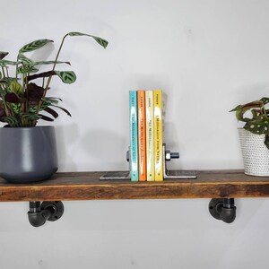 Puede incluir: Una estanter&iacute;a de madera r&uacute;stica con soportes de tuber&iacute;a de metal negro, que presenta un conjunto de libros con un sujetalibros, una planta en maceta con hojas verdes y blancas y una planta en maceta con hojas verdes y marrones.