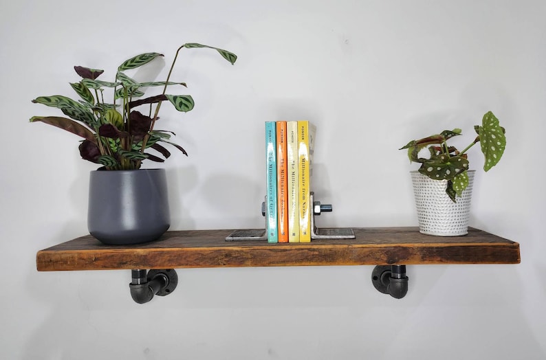 Puede incluir: Una estanter&iacute;a de madera r&uacute;stica con soportes de tuber&iacute;a de metal negro, que presenta un conjunto de libros con un sujetalibros, una planta en maceta con hojas verdes y blancas y una planta en maceta con hojas verdes y marrones.