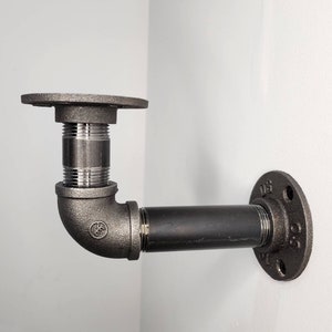 Puede incluir: Soporte de estante de tuber&iacute;a de metal negro con un codo de 90 grados y dos bridas. El soporte est&aacute; montado en una pared blanca.