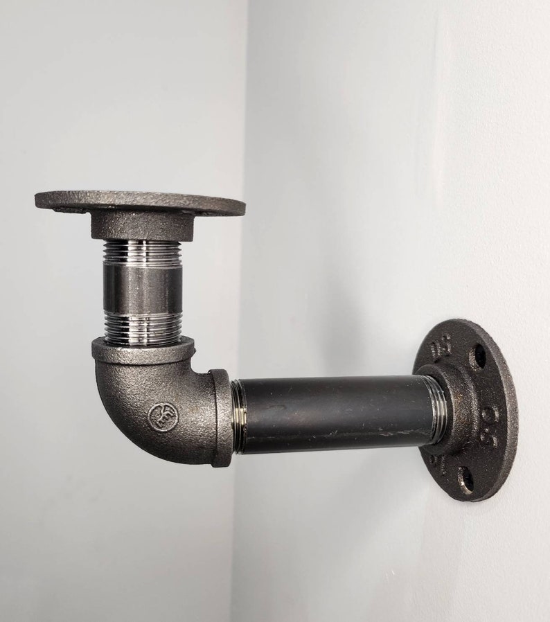 Puede incluir: Soporte de estante de tuber&iacute;a de metal negro con un codo de 90 grados y dos bridas. El soporte est&aacute; montado en una pared blanca.
