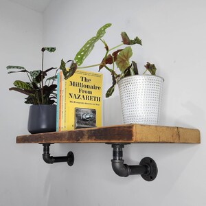 Puede incluir: Una estanter&iacute;a de madera r&uacute;stica con soportes de tuber&iacute;a de metal negro. La estanter&iacute;a est&aacute; decorada con una planta en maceta, un libro titulado "The Millionaire From Nazareth" y una maceta plateada.