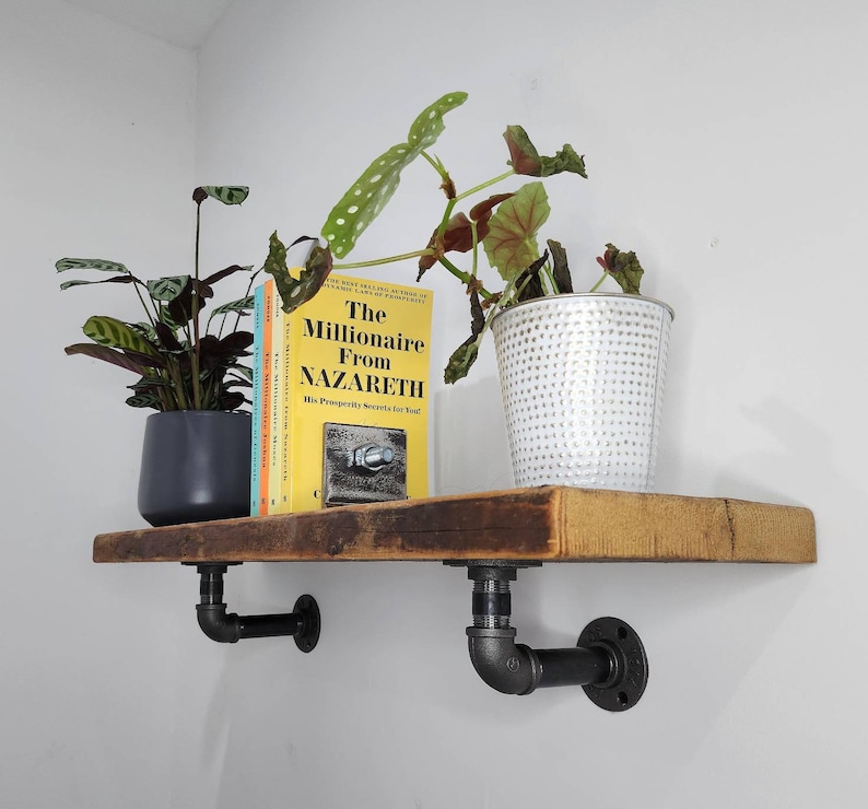 Puede incluir: Una estanter&iacute;a de madera r&uacute;stica con soportes de tuber&iacute;a de metal negro. La estanter&iacute;a est&aacute; decorada con una planta en maceta, un libro titulado "The Millionaire From Nazareth" y una maceta plateada.