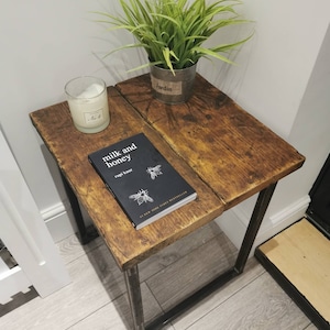 Pode incluir: Uma mesa de apoio de madeira rústica com estrutura de metal preto. A mesa tem um acabamento de madeira envelhecida e é decorada com uma planta em vaso, uma vela e um livro intitulado "milk and honey" de Rupi Kaur.