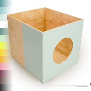 Può includere: Una scatola di legno blu chiaro con un'apertura circolare. La scatola è in compensato e ha una finitura in legno naturale su un lato. La scatola è larga circa 30 cm e alta 30 cm.