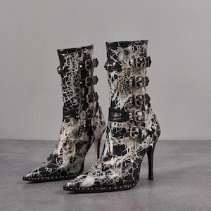 May include: A pair of mid-calf boots featuring a black and white marbled pattern. Each boot has a pointed toe, a high stiletto heel, and four horizontal straps with metal buckles along the side. Small metal studs line the base of the sole.