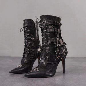 May include: A pair of black leather mid-calf boots featuring pointed toes, high stiletto heels, and front lace-up closures. The boots are adorned with multiple buckled straps, silver-toned metal hardware, and decorative metallic accents on the toes.