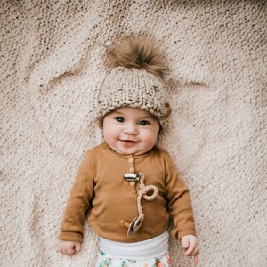 May include: A baby wearing a brown long-sleeved shirt, white pants with a Christmas tree print, and a beige knitted hat with a brown pom-pom.