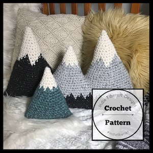May include: Crochet pattern for a mountain-shaped pillow. The pattern is available for purchase and includes instructions for making a pillow with a white peak and a black, gray, or teal base. The pattern is designed by Indie Fiberart Designs.