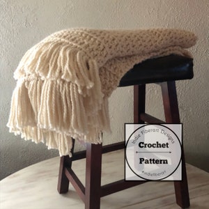 May include: A crocheted throw blanket with fringe in a cream color draped over a wooden stool. The blanket is made with a textured stitch pattern. A white circle with black text reads "Indie Fiberart Designs Crochet Pattern #indiefiberart".
