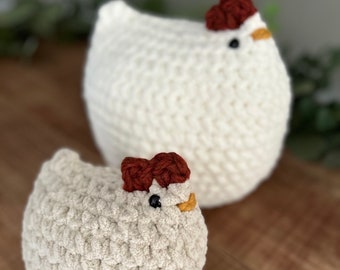 May include: Two crocheted white chickens with brown and yellow accents. The larger chicken is behind the smaller chicken.