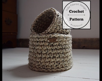 May include: Two crocheted baskets, one nested inside the other, are made with a beige and black speckled yarn. The baskets are sitting on a white wooden floor. A white label with the text "Indie Fiberart Designs Crochet Pattern #indiefiberart" is visible in the top right corner of the image.