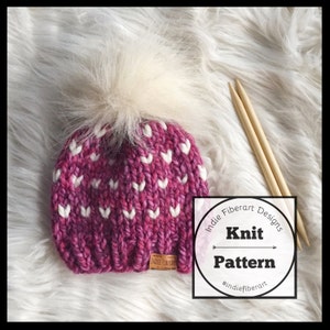 May include: A pink and white knit hat with a white pom pom. The hat has a heart pattern and is accompanied by two wooden knitting needles. The text "Indie Fiberart Designs Knit Pattern #indiefiberart" is on a white circle.