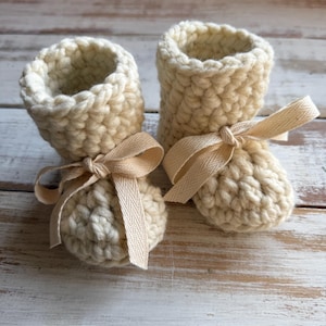 May include: A pair of cream-colored, hand-crocheted baby booties. Each bootie has a rounded toe and a rolled cuff. A cream-colored ribbon is tied around the ankle of each bootie. The booties are made of a chunky yarn and are sitting on a white wooden surface.