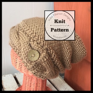Puede incluir: Un gorro de punto marrón, holgado y caído, con un botón grande en un lateral. El gorro está sobre una cabeza de maniquí de mimbre rosa. El texto "Indie Fiberart Designs Knit Pattern #indiefiberart" está en un círculo alrededor del gorro.