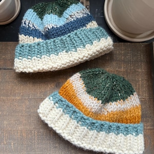 May include: Two knitted hats with colorful stripes. The hats are made with a chunky yarn and have a white brim. The hats are on a wooden surface.
